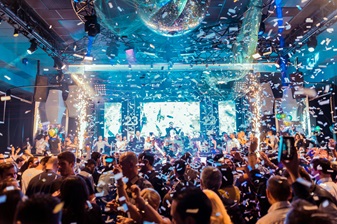 A crowded party with people dancing and cheering under a large disco ball, surrounded by flying confetti and bright stage lights.