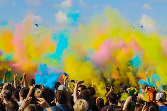 A large crowd of people celebrates with colorful powder in the air at an outdoor event.