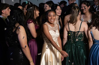 A group of young people in formal attire stand closely together at an event, with some smiling and confetti in the air. The central figure in a cream-colored dress is looking directly at the camera.