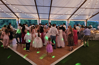 A group of people are dancing under a tent with string lights at a gathering or event. Some are wearing formal attire, and children are also present.